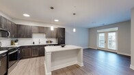 Located at 1420 W Deihl Rd in Naperville, IL, this well-appointed one-bedroom, one-bathroom unit offers a thoughtfully designed open-concept layout. The combined kitchen and living area serves as the heart of the home, showcasing rich dark cabinetry, white subway tile backsplash, light stone countertops, and a central kitchen island with pendant lighting overhead. Stainless steel appliances complete the kitchen, while light-toned hardwood-style flooring extends throughout the main living areas, creating a bright and cohesive aesthetic. The living room benefits from natural light and provides access to a private balcony through elegant French doors, offering views of the surrounding community. The spacious primary bedroom features plush carpeting, a ceiling fan, and a window that invites additional natural light. A dedicated walk-in closet with wire shelving systems provides generous storage. The bathroom is finished with a large glass-enclosed walk-in shower adorned with white subway tile, brushed nickel fixtures, and a dark vanity topped with a marble-style countertop. Neutral tones and clean lines throughout lend the space a modern, move-in-ready appeal.