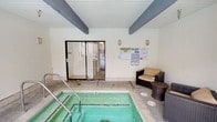 Located at 5825 Reseda Blvd in the Tarzana neighborhood of Los Angeles, this amenity space offers a well-appointed recreational area designed for both relaxation and leisure. The centerpiece is a generously sized outdoor swimming pool with crystal-clear turquoise water, framed by a wide concrete pool deck furnished with modern reclining lounge chairs. Tall, mature trees line the perimeter, providing natural shade and a sense of privacy, while a secure wrought-iron fence encloses the area. A covered walkway runs alongside the pool, supported by painted structural beams and accented with potted greenery, creating a pleasant transitional space between the indoor and outdoor areas. Adjacent to the outdoor pool, an enclosed indoor spa room houses a compact hot tub with handrail entry, set beneath a beamed ceiling with a clean white finish. The spa room includes wall-mounted lighting, a glass sliding door connecting to the pool area, and seating with a wicker sofa and side table, offering a comfortable retreat. Posted pool rules and safety information are prominently displayed throughout. The overall setting reflects a thoughtfully maintained community amenity space suited for year-round use in the Southern California climate.