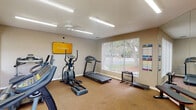 This fitness center is designed to provide a comprehensive workout experience within a single, open floor layout. The room is finished with neutral beige walls and durable carpet flooring, creating a clean and functional environment suited for a variety of exercise routines. Bright fluorescent strip lighting runs across the ceiling alongside a ceiling fan, ensuring the space remains well-lit and comfortable during use. A large window fitted with vertical blinds allows natural light to flow in while offering views of the surrounding greenery outdoors.

The cardio section is well-stocked, featuring multiple treadmills, elliptical cross-trainers, and stationary bikes, accommodating several users simultaneously. Wall-mounted flat-screen televisions are positioned for easy viewing during workouts. For strength training, the space includes a dedicated area with a weight bench, a dumbbell rack, kettlebells, medicine balls in assorted sizes, and resistance tools organized on a freestanding equipment rack. Workout reference charts and instructional posters are displayed along the walls to guide users through exercises. A full-length mirror section reflects the room, adding to the sense of openness. The overall ambiance is practical and motivating, offering everything needed for a complete fitness routine.