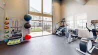 Located in Manteca, California, this fitness center offers a thoughtfully designed exercise space that combines functionality with a bright, welcoming atmosphere. The room features soaring vaulted ceilings with a distinctive wood-paneled finish and recessed lighting, creating an open and airy environment that feels larger than its footprint suggests.

Large floor-to-ceiling windows and a full-width glass garage-style door flood the space with natural light and provide views of the surrounding outdoor areas, including glimpses of a pool and palm trees. The durable dark tile flooring is well-suited for a high-activity environment.

The fitness center is comprehensively equipped for a full-body workout. Cardio options include a StairMaster step climber, an elliptical trainer, and a rowing machine. Strength training is supported by a multi-function cable machine, an adjustable weight bench, a full rack of dumbbells, and a selection of medicine and stability balls neatly stored on racks. A wall-mounted flat-screen television adds entertainment for longer workout sessions, while full-length mirrors along one wall enhance the sense of space and allow for form monitoring. A water station near the entrance ensures convenience. Fitness guidelines are posted at the entry door, contributing to an organized and community-focused environment.