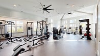 Located in Wildwood, FL, this fitness center offers a thoughtfully designed exercise space that supports a wide range of workout routines. The single-floor layout provides a generous, open floor plan with soaring vaulted ceilings that create an airy, expansive atmosphere. Large windows along multiple walls flood the interior with natural light and offer views of the surrounding outdoor environment, enhancing the overall workout experience.

The space is outfitted with an impressive selection of equipment catering to both strength and cardio training. Strength equipment includes multi-station cable machines, a power rack with bench press, a full dumbbell rack with flat and adjustable benches, and a dedicated free weights area. A large wall mirror runs alongside the free weights section, allowing for proper form checks during training. The cardio zone features treadmills, a stationary bike, and an air bike, while a functional training station stocks kettlebells, medicine balls, a punching bag, and a TKO functional rig. A wall-mounted television provides entertainment during workouts.

Durable flooring throughout and multiple large ceiling fans ensure comfort during intense sessions. Direct outdoor access via glass-panel doors adds convenience. The clean, white and neutral color palette, accented by an inspirational "Keep Growing" wall graphic, creates a motivating and professional environment.