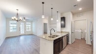 Located in El Paso's Paseo del Este neighborhood, this one-bedroom, one-bathroom residence offers a thoughtfully designed single-floor layout with modern finishes throughout. The open-concept kitchen and living area forms the heart of the home, anchored by a granite-topped kitchen island with a double sink and pendant lighting above. Dark espresso cabinetry contrasts with the light countertops and wood-look vinyl flooring, while stainless steel appliances add a contemporary touch. Built-in shelving provides functional display and storage space adjacent to the kitchen. The living area benefits from natural light through large windows and a glass door that opens to a private outdoor balcony, complemented by a ceiling fan overhead. The bedroom is a comfortable retreat featuring plush carpet flooring, a ceiling fan, and arched doorways leading to both the bathroom and a generously sized walk-in closet outfitted with multi-level shelving and hanging rods. The full bathroom includes a tiled tub-shower combination with oil-rubbed bronze fixtures and wood-look flooring. A dedicated laundry room houses a full-size washer and dryer alongside a water heater, completing this well-appointed residence.