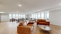 Located at 3425 Eischen Ct in Duluth, Minnesota, this single-floor community space offers a generously sized, open-concept layout designed for comfortable gathering and socializing. The interior is finished with light hardwood-style flooring, recessed lighting, and crisp white walls that give the space a clean, modern aesthetic throughout.

The main lounge area features multiple seating arrangements, including rich cognac leather sofas and loveseats centered around a sleek dark-tiled fireplace with a wall-mounted television — creating a warm and inviting focal point. A separate dining area provides counter-height tables and upholstered stools, ideal for casual meals or conversation. A kitchenette equipped with light wood cabinetry and a stainless steel refrigerator offers convenient amenities for residents and guests.

Floor-to-ceiling windows line the walls, flooding the interior with natural light and framing expansive views of the surrounding Duluth landscape. These windows open visually to a broad wraparound terrace finished with large-format tile pavers and outfitted with Adirondack-style lounge chairs — an inviting outdoor extension of the interior gathering space. The terrace is enclosed by sleek metal railings, offering unobstructed views of the greenery below. Together, the indoor and outdoor areas create a seamless, light-filled retreat suited for both relaxation and community engagement.