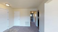 This single-floor space presents a clean, functional layout designed for comfortable everyday living. The main living area is finished with plush carpeting in a neutral gray-brown tone, which extends throughout the hallway and into adjoining rooms, creating a cohesive and understated aesthetic. Smooth, light-colored walls complement the textured ceiling, giving the space a bright and airy feel.

A white interior door provides access to a closet or utility area, while a floor-level ventilation register indicates a built-in HVAC system for year-round climate control. The hallway, illuminated by a flush-mount ceiling fixture, leads toward additional rooms, with a window at the far end drawing in natural light and enhancing the sense of depth throughout the unit.

To one side, a partial view reveals a darker-toned area that appears to transition into a kitchen space, suggesting a practical separation between living and cooking areas. The overall design is straightforward and well-maintained, with white trim and baseboards providing clean definition between walls and floors throughout the space.