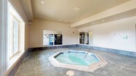 This single-floor indoor recreational space centers around a generously sized octagonal hot tub, finished with blue tile accents and equipped with stainless steel entry rails for easy access. The room is defined by its durable slate-style tile flooring, which extends throughout the entire area and is complemented by a dark tile wainscoting along the lower walls, lending the space a clean, polished appearance.

The walls are finished in a warm neutral tone, and recessed ceiling lighting provides even, ambient illumination throughout. Elevated ceilings contribute to an open, airy atmosphere within the enclosed space. A large window along one wall allows natural light to filter in, offering a view to an outdoor seating area beyond.

Interior windows open to an adjacent vending area, which houses a Pepsi beverage machine and a snack vending unit, providing convenient refreshment access. A mirrored pass-through panel also offers a view into what appears to be a restroom or changing area nearby.

The space features multiple access points, including a set of double glass doors leading to the exterior and an interior access door for building connectivity. An emergency call station is mounted on the wall, reflecting a commitment to safety. Overall, the space is well-designed for guest comfort and convenience in a hospitality environment.