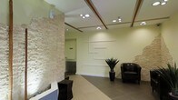 This single-floor space presents a refined and contemporary lobby area designed to make a strong architectural statement. The most striking feature is a prominent natural stacked-stone accent wall, its warm sandy tones providing a striking textural contrast against the smooth, light sage-green painted walls. The ceiling is thoughtfully detailed with exposed wooden beam accents framing recessed circular spotlights, which cast a clean, even illumination throughout the space. The flooring consists of large-format dark grey tiles, grounding the interior with a sense of solidity and sophistication. A geometric wall art installation composed of intersecting rectangular frames adds a modern, sculptural element to the main wall. The seating area features a dark leather armchair alongside a tall, angular side table, offering a comfortable and stylish spot for visitors. A bold potted plant with dramatic, upright foliage serves as a natural focal point, adding a refreshing organic element to the otherwise sleek interior. A small water feature near the stone wall introduces a calming, sensory dimension to the space. Overall, the lobby balances natural materials with clean modern lines, projecting a professional yet inviting atmosphere.