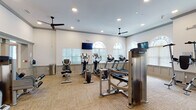 This fitness center offers a thoughtfully designed, single-floor layout that accommodates a full range of workout options. The space is divided into distinct functional zones, providing ample room for both cardio and strength training activities without crowding.

The cardio area is lined with multiple treadmills, elliptical machines, and stationary bikes — including Precor equipment — all positioned in front of arched windows fitted with blinds that allow for natural light while maintaining comfort. Wall-mounted flat-screen televisions are stationed throughout, and ceiling fans help maintain airflow across the workout floor.

The strength training zone features a comprehensive free weights section with a full rack of dumbbells and medicine balls, accompanied by adjustable benches for versatile use. Large floor-to-ceiling mirrors span one wall, enhancing the sense of space and allowing users to monitor their form. A cable machine and additional strength equipment round out this area.

Throughout the facility, wainscoting with a gray-toned finish adds a polished, residential aesthetic to the walls, complemented by neutral toned carpet tiles underfoot. Recessed lighting provides consistent, even illumination across the entire space. The open layout, high ceilings, and generous natural light create a clean, motivating environment well-suited for a variety of fitness routines.