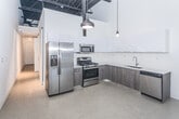 Located at 1209 Morse Ave in Royal Oak, Michigan, this single-floor space showcases a modern industrial aesthetic defined by soaring ceiling heights, polished concrete floors, and a clean, open layout. The main living area makes an immediate impression with its dramatic floor-to-ceiling windows and large glass sliding doors, which flood the interior with natural light and provide direct views of the surrounding streetscape. Exposed ceiling elements and a ceiling fan add an industrial character to the bright, open volume.

The kitchen is thoughtfully designed with an L-shaped configuration featuring two-toned cabinetry — high-gloss white upper cabinets contrast with wood-grain lower cabinets — set against a white backsplash and light countertops. A full suite of stainless steel appliances includes a gas range, built-in microwave, refrigerator, and dishwasher. Pendant lighting adds warmth to the space.

The bathroom presents a clean, minimalist design with large-format tile flooring, a tub-shower combination finished in crisp white tile, a floating vanity, and chrome fixtures throughout. The overall ambiance of the space is contemporary and versatile, offering a bright, well-appointed interior with a strong architectural presence.