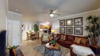 Located at 5799 Southland Dr in Mobile, Alabama, this single-floor residence offers a thoughtfully designed layout that balances comfort and functionality. The open-concept living and dining area serves as the heart of the home, featuring crown molding, wood-look flooring, and a ceiling fan overhead. The living room provides ample space for relaxed seating, accented by a gallery wall arrangement and large windows that invite natural light. Adjacent to the living area, the dining space features wainscoting detail and a dedicated nook framed by decorative wall art. The kitchen is outfitted with warm wood cabinetry, granite-style countertops, stainless steel appliances including a range, microwave, and dishwasher, and a breakfast bar for casual dining. A built-in desk area with a granite countertop offers a convenient workspace. The bedrooms are carpeted and generously sized, each featuring ceiling fans and walk-in closet access. The bathroom includes a single-sink vanity with wood cabinetry, a large mirror with overhead lighting, and a full tub-shower combination with tile surround. Throughout the residence, a cohesive neutral palette and consistent wood-look flooring tie the spaces together, creating a warm and inviting atmosphere.