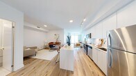 This well-appointed 1-bedroom, 1-bathroom unit offers a thoughtfully designed layout spread across a single floor. The entrance opens into a bright hallway with light wood flooring and a mirrored closet, setting a clean, contemporary tone throughout. The open-concept kitchen and living area form the heart of the unit, featuring a sleek white kitchen with flat-panel cabinetry, white subway tile backsplash, stainless steel appliances including a range, microwave, and refrigerator, and a central island with seating. The living area benefits from floor-to-ceiling windows and a sliding glass door leading to a balcony, flooding the space with natural light and offering sweeping views of the water and city skyline. The bedroom is a serene retreat with a large window framing striking city views, light wood flooring, and a mirrored sliding closet. The bathroom is finished with clean white large-format tiles surrounding a full soaking tub and shower combination. Throughout the unit, warm-toned light wood flooring, recessed and track lighting, and crisp white walls create a cohesive, modern aesthetic with a bright and airy ambiance.