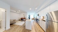 This well-appointed 1-bedroom, 1-bathroom unit offers a thoughtfully designed layout spread across a single floor. The entrance opens into a bright hallway with light wood flooring and a mirrored closet, setting a clean, contemporary tone throughout. The open-concept kitchen and living area form the heart of the unit, featuring a sleek white kitchen with flat-panel cabinetry, white subway tile backsplash, stainless steel appliances including a range, microwave, and refrigerator, and a central island with seating. The living area benefits from floor-to-ceiling windows and a sliding glass door leading to a balcony, flooding the space with natural light and offering sweeping views of the water and city skyline. The bedroom is a serene retreat with a large window framing striking city views, light wood flooring, and a mirrored sliding closet. The bathroom is finished with clean white large-format tiles surrounding a full soaking tub and shower combination. Throughout the unit, warm-toned light wood flooring, recessed and track lighting, and crisp white walls create a cohesive, modern aesthetic with a bright and airy ambiance.