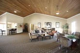 This inviting single-floor space exudes a refined yet comfortable ambiance, defined by its dramatic vaulted ceilings clad in rich, warm-toned wood planking that runs throughout the interior. The centerpiece of the main gathering area is a grand floor-to-ceiling stone fireplace, crafted from natural stacked stone, which anchors the space and lends a rustic yet sophisticated character. Recessed lighting and ceiling fans complement the natural light filtering in through multiple windows fitted with slatted blinds.

The patterned carpet — featuring a flowing dark scrollwork design — flows seamlessly across the entire floor, tying the various seating areas together cohesively. Multiple conversational arrangements are thoughtfully distributed throughout, featuring wicker and upholstered chairs, side tables, and accent lamps. The walls are adorned with a curated collection of framed artwork, adding visual interest and a cultured atmosphere.

A dedicated dining area accommodates guests around a glass-topped table with elegantly carved wooden chairs, positioned beside large windows that allow for an airy, light-filled dining experience. A framed vintage-style poster serves as a focal point on the dining room wall. Throughout the space, decorative accents including potted plants and sculpted vessels contribute to a warm, welcoming environment well-suited for gatherings and relaxation.