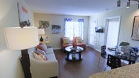 This single-floor space offers a warm and inviting atmosphere anchored by rich hardwood-style flooring that runs throughout the main living area. The living room is generously proportioned, featuring a comfortable neutral sofa adorned with colorful patterned throw pillows, a pair of rust-toned accent chairs, and a round dark wood coffee table that serves as a natural focal point for the seating arrangement. Tropical-themed wall art adds a playful, relaxed character to the space, complementing the cheerful color palette of warm oranges, yellows, and greens. Natural light floods the room through large windows dressed with bold blue-and-white chevron curtains and classic white plantation shutters, creating a bright and airy feel. A wall-mounted flat-screen television is positioned conveniently near a secondary window, while a compact dining area with a sleek black chair is tucked neatly to one side, making efficient use of the available space. Smooth walls with a subtle textured ceiling complete the interior finish. The layout flows openly and efficiently, making the space feel both functional and comfortable for everyday living.