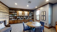 This thoughtfully designed single-floor space blends rustic charm with refined comfort, creating an inviting library and lounge environment. A striking reclaimed wood accent wall serves as a dramatic focal point, adorned with framed artwork and decorative pieces that add character and visual interest. Complementing this feature, a full wall of rich, dark-stained wooden built-in bookshelves lines one side of the room, filled with books and accented by illuminated framed art panels, adding warmth and a scholarly ambiance.

The seating areas are carefully arranged to encourage both intimate conversation and quiet reading. A round wooden table surrounded by navy blue upholstered chairs provides a central gathering spot, while pairs of leather armchairs and tufted wingback chairs are positioned throughout for relaxed lounging. Patterned carpeting in soft, neutral tones anchors each seating arrangement and adds a layer of texture to the space.

Natural light filters in through multiple windows with wood-trimmed frames, offering views of the surrounding greenery. A set of glass-paneled double doors with rich wood framing opens to a grand entry foyer beyond, visible through the panes and featuring a chandelier and stone accents. The space is unified by consistent use of wood detailing, crown molding, and recessed lighting, creating a cohesive and welcoming atmosphere throughout.