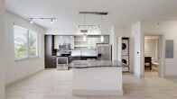 This single-floor residence at 7941 West Dr in North Bay Village, Florida, presents a clean, modern aesthetic throughout its well-appointed layout. The open-concept kitchen serves as a central highlight, featuring a striking combination of dark wood and light cabinetry, granite countertops, a full suite of stainless steel appliances, and a white tile backsplash. A central island with a granite surface provides additional workspace and casual seating. Pendant and track lighting illuminate the kitchen, while a stacked washer and dryer unit is conveniently tucked nearby. The living area flows naturally from the kitchen and is defined by decorative arched architectural details that lend a distinctive character to the space. Large-format light tile flooring runs throughout, creating a cohesive and airy atmosphere. Ceiling fans in the living areas promote comfortable airflow, and windows invite natural light and views of the surrounding tropical greenery. The bathroom is finished with a dark wood vanity topped with granite, a framed mirror, and a tiled shower-tub combination, maintaining the consistent modern design language found throughout the home. Overall, the space offers a bright, functional, and stylish environment in a desirable South Florida island community.