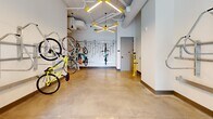 This thoughtfully designed amenity space serves as a dedicated bike storage and resident lounge, offering a well-organized and visually engaging environment across a single floor. The facility is divided into two distinct areas that work seamlessly together.

The bike storage area is outfitted with multiple wall-mounted bicycle racks, accommodating a variety of bike styles and sizes both horizontally and vertically. Polished concrete floors provide a durable, easy-to-maintain surface, while exposed ductwork and pendant lighting with bold yellow accents lend an industrial-modern aesthetic to the space. Large glass entry doors flood the room with natural light and offer views to the outdoor surroundings, creating an open and welcoming atmosphere.

The adjacent lounge area makes a striking visual statement with a warm wood-paneled accent wall featuring illuminated "Live With Grace" lettering. A built-in counter beneath the wall is lined with vibrant blue stools, creating a casual seating nook ideal for socializing or unwinding. Geometric pendant light fixtures with yellow detailing echo the design language found throughout the space. Together, both areas reflect a cohesive, contemporary design philosophy that balances functionality with personality, making this an inviting and practical amenity for cycling enthusiasts and residents alike.