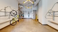 This thoughtfully designed amenity space serves as a dedicated bike storage and resident lounge, offering a well-organized and visually engaging environment across a single floor. The facility is divided into two distinct areas that work seamlessly together.

The bike storage area is outfitted with multiple wall-mounted bicycle racks, accommodating a variety of bike styles and sizes both horizontally and vertically. Polished concrete floors provide a durable, easy-to-maintain surface, while exposed ductwork and pendant lighting with bold yellow accents lend an industrial-modern aesthetic to the space. Large glass entry doors flood the room with natural light and offer views to the outdoor surroundings, creating an open and welcoming atmosphere.

The adjacent lounge area makes a striking visual statement with a warm wood-paneled accent wall featuring illuminated "Live With Grace" lettering. A built-in counter beneath the wall is lined with vibrant blue stools, creating a casual seating nook ideal for socializing or unwinding. Geometric pendant light fixtures with yellow detailing echo the design language found throughout the space. Together, both areas reflect a cohesive, contemporary design philosophy that balances functionality with personality, making this an inviting and practical amenity for cycling enthusiasts and residents alike.