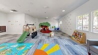 This cheerful, single-floor children's play space is thoughtfully designed to foster creativity, learning, and active play. The open layout provides generous room for children to move freely, with durable, patterned carpet tile flooring in cool grey and blue tones running throughout the entire space.

The room is equipped with a variety of engaging play structures, including a treehouse climber, a safari-themed jeep with a slide, and a colorful caterpillar ride-on toy, offering diverse play experiences for young children. Colorful foam play mats are spread across the floor, providing safe, cushioned areas for floor play.

Educational elements are woven throughout the design, with alphabet and number decals adorning the bright white walls, creating a stimulating learning environment. A dedicated activity area features a child-sized table with bright blue chairs, ideal for arts, crafts, and structured learning activities. A bookshelf stocked with children's books adds a quiet reading corner to the space.

Natural light filters in through multiple windows along the walls, keeping the room airy and inviting. Comfortable grey sofas are positioned throughout, offering caregivers and supervising adults convenient seating with clear sightlines across the room. Recessed and flush-mount ceiling lighting ensures the space remains well-lit at all times. A dedicated entry door provides easy access to and from the play area.