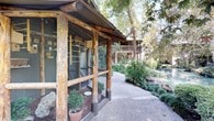 This distinctive single-story space is defined by its deep connection to the natural environment, offering a truly immersive outdoor experience. The exterior features rustic log-framed enclosures with wire mesh paneling, evoking a charming, naturalistic aesthetic that blends seamlessly into the surrounding landscape. Weathered wooden posts and structural elements lend the space a rugged, organic character that feels both timeless and grounded.

A winding concrete pathway guides visitors through the grounds, bordered by abundant greenery, manicured shrubs, and mature deciduous trees that provide generous shade. Potted plants and native vegetation add layers of texture and life throughout the walkways. A standout feature of the exterior is a small decorative pond with a wooden footbridge, its calm green water reflecting the surrounding foliage and contributing to the peaceful, resort-like ambiance.

Adjacent multi-story structures with wood-clad balconies and stairways are visible within the complex, suggesting a cohesive architectural style that prioritizes natural materials and harmony with the landscape. The overall setting feels secluded and tranquil, offering a retreat-like quality rarely found in urban environments. The combination of rustic construction, lush plantings, and water features makes this a uniquely characterful and visually engaging space.