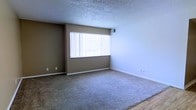 This single-story space presents a straightforward and functional layout with a focus on simplicity. The main living area is finished with wall-to-wall carpet in a neutral beige tone, providing a comfortable and cohesive foundation for the room. Two-toned walls — a warm tan accent wall paired with crisp white side walls — create a subtle visual contrast that adds modest dimension to the interior.

A large picture window fitted with vertical blinds serves as the room's primary source of natural light, brightening the space and offering a connection to the outdoors. The window's prominent size ensures the room feels open and well-lit throughout the day. An overhead HVAC vent is visible near the ceiling, indicating a centralized climate control system.

The ceiling features a textured popcorn finish, a characteristic detail of the space, while clean white baseboards run along the perimeter of the walls, providing a polished border between the carpet and walls. A small section of hard flooring is visible at the edge of the room, suggesting a transition to an adjacent area. Overall, the space maintains a clean, move-in-ready appearance.