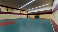 This single-level indoor sports facility is designed to accommodate a variety of athletic activities, centered around a full-size basketball court and additional racquetball courts. The main gymnasium features a spacious, high-ceilinged hall with a tiled sport-surface floor in teal and red, clearly marked with regulation court lines. A regulation basketball hoop is mounted on one end, accompanied by a padded green wall protector beneath it for player safety. The walls are finished in a two-tone yellow and deep burgundy color scheme, adorned with silhouette murals of athletes in various sports poses, adding an energetic and motivational character to the space. Overhead, rows of fluorescent lighting provide consistent, bright illumination across the full extent of the court. The racquetball courts offer a distinctly different atmosphere — clean, enclosed spaces with smooth white walls and polished hardwood flooring marked with red boundary lines. These courts are equipped with overhead lighting and climate control venting, ensuring comfortable playing conditions. Together, the facility presents a well-equipped, purpose-built environment suited for basketball, racquetball, and general athletic use.