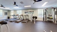 Located at 98 Paddock Club Dr in Panama City Beach, Florida, this dedicated fitness center offers a well-appointed exercise room designed to support a comprehensive workout routine. The space is finished with durable rubber flooring throughout, providing a safe and functional surface for all types of training activities.

The room is generously equipped with a row of treadmills and Matrix elliptical stair climbers for cardiovascular exercise, complemented by a cable/multi-use strength machine, a rowing machine, a dumbbell rack, and stability balls for functional training. Full-length mirrors line one wall, visually expanding the space and allowing users to monitor their form during workouts.

Natural light filters in through multiple white-framed windows fitted with blinds, creating a bright and inviting atmosphere. Ceiling fans are installed throughout, ensuring comfortable air circulation during intense workouts. Recessed and flush-mounted fluorescent lighting provides consistent illumination across the entire room.

A wall-mounted flat-screen television adds entertainment value for cardio sessions, while framed artwork and fitness-related signage contribute to the motivating environment. The clean, light sage-green walls and white trim lend a fresh, polished aesthetic to the space. A dedicated entry door provides direct access to the fitness center, making it a convenient and well-organized amenity.
