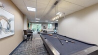 Located at 637 Main St NW in Elk River, Minnesota, this single-floor community amenity space offers a generously sized, open-plan layout designed to accommodate a variety of social and recreational activities. The interior is finished with a drop panel ceiling fitted with recessed lighting throughout, while multi-toned, patterned carpet tiles add visual warmth and texture underfoot.

The space is organized into several distinct functional zones. A fully equipped kitchen anchors one end of the floor plan, featuring dark cabinetry, stainless steel appliances including a refrigerator, microwave, and range, and an island with bar seating — ideal for entertaining or casual gatherings. Adjacent to the kitchen, a comfortable lounge area offers an arrangement of sofas and accent chairs centered around coffee tables, creating a relaxed setting for conversation.

Large windows and glass doors along one wall flood the lounge with natural light and provide views of the surrounding greenery, lending the space an open and airy feel. A game zone features a full-size foosball table and a pool table, catering to leisure and recreational use. Additional seating vignettes include high-top tables with bar-height chairs and a media console with a wall-mounted flat-screen TV. A dark-toned shelving unit serves as a stylish room divider, while a dedicated dining area with pendant lighting and vibrant yellow chairs adds a pop of color.