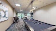 Located at 637 Main St NW in Elk River, Minnesota, this single-floor community amenity space offers a generously sized, open-plan layout designed to accommodate a variety of social and recreational activities. The interior is finished with a drop panel ceiling fitted with recessed lighting throughout, while multi-toned, patterned carpet tiles add visual warmth and texture underfoot.

The space is organized into several distinct functional zones. A fully equipped kitchen anchors one end of the floor plan, featuring dark cabinetry, stainless steel appliances including a refrigerator, microwave, and range, and an island with bar seating — ideal for entertaining or casual gatherings. Adjacent to the kitchen, a comfortable lounge area offers an arrangement of sofas and accent chairs centered around coffee tables, creating a relaxed setting for conversation.

Large windows and glass doors along one wall flood the lounge with natural light and provide views of the surrounding greenery, lending the space an open and airy feel. A game zone features a full-size foosball table and a pool table, catering to leisure and recreational use. Additional seating vignettes include high-top tables with bar-height chairs and a media console with a wall-mounted flat-screen TV. A dark-toned shelving unit serves as a stylish room divider, while a dedicated dining area with pendant lighting and vibrant yellow chairs adds a pop of color.