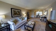 This single-floor residence offers an open-concept layout that seamlessly connects the living and dining areas, creating a cohesive and functional living environment. The living room is finished with wall-to-wall carpeting in a warm beige tone, providing a comfortable foundation for the space. Walls are painted in soft, neutral hues that contribute to the overall inviting ambiance. A textured popcorn ceiling runs throughout, adding a classic touch overhead.

The living area is spacious enough to accommodate a full sofa set alongside a glass-topped coffee table, while a freestanding display shelf provides additional storage and decorative opportunity. A large framed artwork anchors the main wall, lending the room a polished, curated feel.

The dining area is positioned toward the rear of the space, benefiting from natural light filtered through a window. A ceiling fan with light fixture serves this zone, offering both ventilation and illumination. The dining setup comfortably seats multiple guests, making it well-suited for everyday use and casual entertaining. Together, the two areas form a well-balanced, practical living space with a warm, neutral aesthetic throughout.