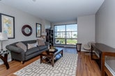 This single-floor residence is thoughtfully designed to balance modern functionality with comfortable living. The kitchen serves as a central hub, outfitted with rich dark-wood cabinetry, stainless steel appliances including a refrigerator, range, built-in microwave, and dishwasher, and light-toned countertops. A central island with a double sink provides additional workspace, while pendant lighting overhead adds a warm, refined touch. Hardwood flooring flows seamlessly from the kitchen into the living area, where large floor-to-ceiling windows flood the space with natural light and frame views of surrounding greenery and neighboring buildings. The living room offers a welcoming atmosphere, anchored by warm-toned hardwood floors and complemented by decorative wall art and open shelving. The bedroom features plush carpeting, soft neutral tones, and windows that allow natural light to filter in, creating a calm and restful environment. The bathroom is finished with tile flooring, a vanity with rich wood cabinetry, a mirrored medicine cabinet, and a well-lit fixture above. Overall, the residence presents a cohesive, contemporary aesthetic with quality finishes throughout.