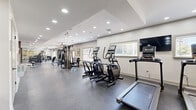 Located at 127 Andorra Glen Ct in Lafayette Hill, PA, this well-equipped fitness center offers a generous, open single-floor layout designed to accommodate a full range of workout activities. The space features durable speckled rubber flooring throughout, providing both safety and comfort underfoot during exercise.

The cardio zone is prominently stocked with multiple treadmills, elliptical machines, and a stair climber, all arranged to maximize floor space and usability. A wall-mounted flat-screen television is positioned in front of the cardio equipment, enhancing the workout experience. The strength training area includes a multi-station cable machine, a dedicated free weights section with a full rack of dumbbells, adjustable benches, a barbell rack, and additional resistance machines. A mirrored wall panel in the free weights area allows for proper form monitoring during lifts.

The ceiling features a coffered design with recessed lighting that evenly illuminates the entire space, while ceiling fans promote air circulation. Multiple windows fitted with blinds invite natural light and offer views of the surrounding greenery. Framed botanical artwork on the neutral-toned walls adds a subtle decorative touch, contributing to a clean and welcoming atmosphere. A glass-paneled entry door provides convenient outdoor access, completing this fully equipped community fitness facility.