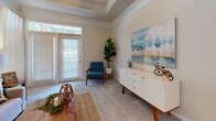 This single-floor residence offers a thoughtfully designed layout with refined finishes throughout. The living room is a standout space, defined by an elegant tray ceiling with crown molding and a ceiling fan. Plush carpet underfoot and a full-light exterior door allow natural light to pour in, creating a bright and welcoming atmosphere with access to the outdoors. The adjacent dining area continues the classic aesthetic with wainscoting panels, crown molding, and a decorative chandelier, all complemented by warm hardwood flooring near the entryway. A round glass-top dining table sits beneath the chandelier, making it an inviting space for meals and gatherings. The kitchen features light wood cabinetry, granite countertops, hardwood flooring, and black appliances including a refrigerator, range, and over-the-range microwave. A window above the counter brings in natural light. The bedroom is a serene retreat with plush carpeting, neutral walls, and a generously sized window dressed with sheer curtains that diffuse soft natural light. The full bathroom offers a long dual-sink vanity with warm wood cabinetry, a white countertop, a large arched mirror, and tile flooring, providing both style and practicality throughout.