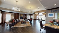 This single-floor community space is thoughtfully designed to blend comfort and functionality across several distinct yet interconnected areas. The main lounge serves as the centerpiece, featuring dark hardwood-style tile flooring, bold crown molding, and a striking stone-surround fireplace flanked by rich wood cabinetry. Plush leather seating and an ornate area rug anchor the space, while a combination of pendant drum fixtures and recessed lighting creates a warm, inviting ambiance. Tray and vaulted ceiling details add architectural interest throughout.

Natural light plays a prominent role, particularly near the dining area, where a dramatic arched window overlooks an outdoor pool area. Multiple large windows dressed with neutral drapes allow daylight to flow generously into the space.

A well-appointed kitchenette offers warm wood cabinetry, granite-style countertops, a mosaic tile backsplash, and full appliances—well-suited for gatherings and events. Adjacent dining areas with round tables and leather chairs provide comfortable seating for small groups.

The entry area features mosaic tile column accents, additional seating, and display areas, creating a welcoming transition into the broader space. Overall, the design balances traditional warmth with practical amenities.
