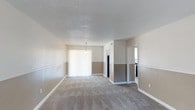 Located at 190 N Corry Field Rd in Pensacola, Florida, this single-floor property offers a clean and functional layout designed for comfortable living. The interior features a warm, neutral two-tone color palette with a contrasting wall treatment that adds a subtle decorative element throughout the main living areas. Soft wall-to-wall carpeting runs through the open living and dining space, providing a comfortable underfoot feel. A textured ceiling adds dimension to the rooms, while a single pendant light fixture anchors the dining area, lending a simple yet defined sense of purpose to the space. Large vertical blinds on a sliding glass door allow for natural light control and provide access to the outdoors. The open floor plan creates a seamless flow between the living and dining areas, with a partial view into what appears to be a kitchen equipped with standard appliances. Doorways leading to additional rooms suggest a well-organized layout. The overall ambiance is neutral, move-in ready, and well-suited for a range of furnishing styles, making it a versatile and practical living space in the Pensacola area.