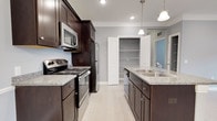 This single-floor space showcases a thoughtfully designed kitchen that serves as its centerpiece. Rich, dark espresso cabinetry lines the walls, providing ample storage and creating a striking contrast against the light gray walls and neutral tile flooring. The kitchen is outfitted with stainless steel appliances, including a refrigerator, over-the-range microwave, and electric range, all complementing the light granite countertops that offer generous prep space. A central island features a double-basin stainless steel sink and additional cabinetry beneath, enhancing both functionality and workflow. Elegant pendant lights hang overhead, adding a refined touch to the space, while recessed lighting ensures the area is well-lit throughout. A dedicated pantry with open shelving provides excellent storage capacity and keeps the kitchen organized and clutter-free. The light-toned tile flooring runs seamlessly underfoot, tying the design elements together. The overall ambiance is clean, modern, and highly functional, with a careful balance of dark and light tones that creates a welcoming and practical environment suited for everyday living and entertaining alike.