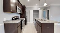 This single-floor space showcases a thoughtfully designed kitchen that serves as its centerpiece. Rich, dark espresso cabinetry lines the walls, providing ample storage and creating a striking contrast against the light gray walls and neutral tile flooring. The kitchen is outfitted with stainless steel appliances, including a refrigerator, over-the-range microwave, and electric range, all complementing the light granite countertops that offer generous prep space. A central island features a double-basin stainless steel sink and additional cabinetry beneath, enhancing both functionality and workflow. Elegant pendant lights hang overhead, adding a refined touch to the space, while recessed lighting ensures the area is well-lit throughout. A dedicated pantry with open shelving provides excellent storage capacity and keeps the kitchen organized and clutter-free. The light-toned tile flooring runs seamlessly underfoot, tying the design elements together. The overall ambiance is clean, modern, and highly functional, with a careful balance of dark and light tones that creates a welcoming and practical environment suited for everyday living and entertaining alike.