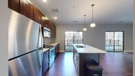 Located at 245 S High St in the heart of downtown Columbus, Ohio, this one-bedroom, one-bathroom residence offers a well-designed layout that maximizes every square foot of its efficient floor plan. The combined kitchen and living area serves as the centerpiece of the unit, featuring rich dark-stained cabinetry extending to the ceiling, quartz countertops, a subway tile backsplash, and a full suite of stainless steel appliances including a refrigerator, electric range, and built-in microwave. A large kitchen island with a undermount sink provides additional prep space and casual seating. Sleek pendant lighting illuminates the island, while hardwood-style flooring runs throughout the main living areas, adding warmth and continuity. Black-framed sliding glass doors lead to a private balcony overlooking the surrounding downtown Columbus streetscape, allowing abundant natural light to fill the living room. The bedroom is carpeted for comfort and includes a walk-in closet with wire shelving. The bathroom is finished with a wood-toned vanity and white countertop. Additional conveniences include an in-unit laundry area and a hallway connecting the private spaces. The overall aesthetic is contemporary urban, with neutral gray walls and modern finishes throughout.