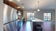 Located at 245 S High St in the heart of downtown Columbus, Ohio, this one-bedroom, one-bathroom residence offers a well-designed layout that maximizes every square foot of its efficient floor plan. The combined kitchen and living area serves as the centerpiece of the unit, featuring rich dark-stained cabinetry extending to the ceiling, quartz countertops, a subway tile backsplash, and a full suite of stainless steel appliances including a refrigerator, electric range, and built-in microwave. A large kitchen island with a undermount sink provides additional prep space and casual seating. Sleek pendant lighting illuminates the island, while hardwood-style flooring runs throughout the main living areas, adding warmth and continuity. Black-framed sliding glass doors lead to a private balcony overlooking the surrounding downtown Columbus streetscape, allowing abundant natural light to fill the living room. The bedroom is carpeted for comfort and includes a walk-in closet with wire shelving. The bathroom is finished with a wood-toned vanity and white countertop. Additional conveniences include an in-unit laundry area and a hallway connecting the private spaces. The overall aesthetic is contemporary urban, with neutral gray walls and modern finishes throughout.