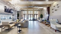 This expansive single-floor clubhouse is a masterfully designed communal amenity space that blends contemporary elegance with inviting comfort. Soaring ceilings with exposed wood beams set a dramatic architectural tone throughout, while large-format tile flooring in warm neutral tones flows seamlessly across the open-plan layout. The main lounge area is anchored by a striking floor-to-ceiling accent wall clad in textured tile, framing a sleek linear fireplace beneath a wall-mounted television. A statement sputnik chandelier adds a sculptural focal point overhead. Floor-to-ceiling windows and sets of French doors flood the interior with abundant natural light and offer direct views of an outdoor pool area, creating a seamless indoor-outdoor connection. The space transitions into a formal dining zone with a dark-wood dining table set beneath exposed beam ceilings. A fully equipped catering kitchen features rich dark cabinetry, a gleaming mosaic tile backsplash, stainless steel appliances, and an oversized granite island with bar seating illuminated by globe pendant lights. A game room area with a shuffleboard table and additional bar counter rounds out the amenities. Multiple lounge seating arrangements with contemporary furnishings provide versatile spaces for relaxation and socializing throughout.