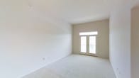 This single-floor space presents a clean, well-lit interior defined by a neutral palette and an open, airy atmosphere. The main room is immediately notable for its elevated ceilings, which enhance the sense of volume and openness throughout the space. Soft, light-toned wall-to-wall carpeting lines the floor, adding warmth and comfort underfoot while complementing the crisp white walls.

A standout architectural feature is the set of French doors positioned at the far end of the room, framed by a transom window above. This combination allows an abundance of natural light to pour in, brightening the interior and creating a seamless visual connection to the outdoors. The doors suggest access to a balcony or exterior area, further extending the livable feel of the space.

The walls are finished in a clean, neutral beige-white tone that reflects light effectively and provides a versatile backdrop. The overall design is simple and understated, emphasizing functionality and adaptability. The space is presented in a vacant state, offering a clear view of its architectural characteristics without the visual interruption of furnishings. The high ceilings, plentiful natural light, and well-proportioned layout make this a bright and inviting environment with strong potential for a variety of uses.