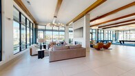 Located at 2200 Cascade Pointe Blvd in Charlotte, NC, this impressive single-floor communal living space offers a generously sized, open-concept layout designed for both relaxation and social interaction. The interior is anchored by striking exposed wood ceiling beams that contrast beautifully against the bright white ceiling, lending the space a modern yet warm architectural character. Large-format white tile flooring flows seamlessly throughout, enhancing the sense of openness and continuity.

Floor-to-ceiling black-framed windows line the perimeter, flooding the interior with abundant natural light and offering panoramic views of the surrounding Cascade Pointe community. The main lounge area features plush sectional sofas, accent chairs, and a sculptural globe chandelier, all centered around a sleek fireplace with a tile surround and mounted flat-screen television. A bold navy-blue area rug defines the game room zone, which includes a billiards table and bar-height seating along a media wall.

Additional seating vignettes are dispersed throughout, offering intimate gathering spots accented by vibrant teal and gold tones. Curated wall art, built-in bench seating, and decorative pendant lighting add refined detail throughout. Glass doors open directly to an outdoor terrace, further connecting the interior to the surrounding environment. The overall ambiance is contemporary, vibrant, and inviting.