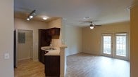 This single-floor residence offers a well-designed open-concept layout that connects the kitchen and living area into one cohesive, functional space. The kitchen is appointed with rich dark wood cabinetry, a neutral tile backsplash, and a light-toned countertop that extends into a breakfast bar peninsula, providing both additional workspace and casual seating. A stainless steel sink is neatly integrated into the counter, while recessed and flush-mount ceiling lighting ensures the kitchen remains bright and practical.

The transition into the living area is defined by a decorative column, adding a subtle architectural detail that helps delineate the two spaces without sacrificing openness. Light wood-toned flooring runs throughout, contributing warmth and visual continuity across the entire floor plan. The living area is equipped with a ceiling fan and light fixture, promoting comfortable air circulation year-round.

Natural light filters into the space through a glass-paneled door along the far wall, offering both a view to the exterior and convenient outdoor access. The overall ambiance is clean, neutral, and inviting, making this a highly functional and comfortable single-floor living space.