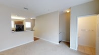 This two-floor residence offers a functional and comfortable layout designed for everyday living. The main level features an open living area finished with plush wall-to-wall carpeting, creating a warm and inviting atmosphere throughout. Neutral beige walls complement the light-toned flooring, lending the space a clean and cohesive aesthetic. A kitchen area is visible from the main living space, highlighted by dark countertops that provide a striking contrast to the surrounding light palette. The kitchen is equipped with appliances and benefits from natural light streaming in through a nearby window. A staircase with a white railing connects the two floors, facilitating easy movement between levels and adding a subtle architectural element to the interior. The upper level continues the consistent design language established on the main floor. Recessed lighting and ceiling-mounted fixtures provide ample illumination throughout, ensuring a bright and well-lit environment. Crisp white trim and baseboards throughout offer a polished finishing touch. The overall ambiance is modest yet well-maintained, making this a practical and livable space suited to a variety of lifestyles.