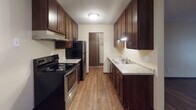 Located at 1430 100th Ave NW in Coon Rapids, Minnesota, this single-floor unit offers a thoughtfully laid-out living space with a consistent, clean aesthetic throughout. The living area is generously sized, featuring dark hardwood-style flooring, neutral-toned walls, and a textured ceiling that adds subtle dimension. A large sliding glass door fitted with vertical blinds fills the room with natural light and provides views of the surrounding outdoor greenery, while a wall-mounted air conditioning unit and baseboard heating ensure year-round comfort. The galley-style kitchen is well-appointed, showcasing rich dark wood cabinetry lining both walls, light-toned countertops, a stainless steel sink, and black appliances including a range and refrigerator. A doorway at the end of the kitchen leads to a dedicated storage or pantry area with built-in shelving. The bathroom features earthy slate-look tile flooring, dark wood vanity cabinetry with brushed nickel hardware, a white countertop, and a full tub-shower combination with tiled surround. A walk-in closet with built-in shelving and hanging rods provides ample storage. The overall layout flows efficiently, making practical use of every area throughout the home.