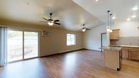 This single-floor space features an expansive open-concept layout that seamlessly connects the living area to the kitchen. The living area is finished with warm-toned hardwood flooring that runs throughout, lending a clean and cohesive feel to the interior. Two ceiling fans are installed across the space, providing ample air circulation and contributing to the room's functional design.

The ceilings are notably varied, with a raised tray ceiling section in part of the living area adding architectural interest and a sense of added height. Natural light is introduced through multiple windows, including a large sliding glass door that opens to a balcony, offering outdoor views and additional ventilation. A wall-mounted air conditioning unit is also present near the balcony entrance.

Baseboard heating runs along the walls, offering a practical climate control solution. The kitchen, partially visible from the main living area, features light wood cabinetry and granite-style countertops, complemented by a pendant light fixture overhead. The neutral beige wall tones throughout the space provide a versatile backdrop, making it well-suited for a variety of interior styles.
