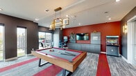 Located at 269 S State Highway 360 in Mansfield, Texas, this single-floor recreational and lounge space is designed to offer an engaging, social environment with a bold, modern aesthetic. The room's design centers on a full-size pool table positioned beneath a striking geometric gold pendant light fixture, which serves as the visual centerpiece of the space. The flooring features a dynamic patterned carpet with bold red accent stripes that guide the eye throughout the room.

A deep crimson feature wall anchors the entertainment side of the room, where a wall-mounted flat-screen TV is flanked by built-in grey cabinetry with open shelving for display. Alongside the cabinetry, a classic arcade game cabinet adds a retro gaming element that complements the space's recreational theme. The walls throughout are finished in a sophisticated dark charcoal grey, contributing to the energetic yet polished atmosphere.

Adjacent to the main game area, a comfortable lounge nook offers cushioned bench seating and bistro-style tables, ideal for casual conversation or spectating. Floor-to-ceiling windows and large sliding glass panels flood the space with natural light while providing views of the surrounding exterior grounds. Gold wall sconces add warm accent lighting, enhancing the contemporary design. French doors connect the game room to a neighboring lounge area, making the overall layout open and socially fluid.