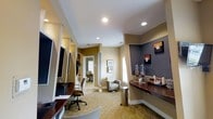 This single-floor professional office space is thoughtfully designed to provide a functional and welcoming environment for both staff and visitors. The reception and waiting area serves as the heart of the space, featuring warm neutral tones — including taupe, beige, and slate — across the walls, creating a calm and professional atmosphere. The patterned carpet adds texture and visual interest underfoot, while recessed ceiling lighting provides consistent, even illumination throughout.

A beverage station along one wall is equipped with drink dispensers, cups, and counter space atop a rich, dark granite-style surface, offering convenience for guests. Adjacent to this area, a compact lounge seating arrangement includes an upholstered armchair and a small side table positioned near a window, allowing natural light to filter into the space.

Workstations with ergonomic office chairs and large flat-screen monitors are integrated along a built-in dark-countered desk area, supporting staff productivity. A corridor leads further into the space, revealing additional private offices furnished with task chairs and desks. Framed artwork adorns the walls, adding a polished, professional touch to the overall design.