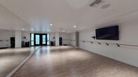 Located on Hollywood Boulevard in Los Angeles, this single-floor studio is thoughtfully designed to support dance, barre, and fitness instruction. The open layout provides generous, unobstructed floor space finished with smooth light wood flooring ideal for movement and exercise. Floor-to-ceiling mirrors line multiple walls, creating a sense of expanded space while allowing practitioners to monitor form and technique. Wall-mounted ballet barres run the length of the mirrored and adjacent walls, catering to barre classes and dance training. A large flat-screen television is mounted prominently on one wall, supporting instructional or media-driven workouts. Recessed lighting throughout the ceiling ensures the space is consistently and evenly illuminated. Black-framed double glass doors provide access to the exterior, allowing natural light to filter in and offering a clean, modern aesthetic. The white walls and minimalist design give the studio a bright, professional ambiance. Additional equipment storage shelving is positioned near the entrance. The overall environment is clean, functional, and well-suited for a variety of movement-based disciplines in the heart of Hollywood.