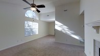 This single-story home is designed to feel open and welcoming, with vaulted ceilings that lend a sense of generous vertical space throughout the main living areas. The living room serves as a central gathering space, highlighted by a classic tiled fireplace with a clean white mantel — an ideal focal point for the room. Plush wall-to-wall carpeting runs throughout, adding warmth and comfort underfoot. Ceiling fans are installed in multiple rooms, providing both functional airflow and a practical design touch. Natural light is a defining feature of this home, with a striking arched window positioned high on the wall allowing sunlight to flood into the space at multiple angles, casting bright, sweeping light across the interior. Standard windows with blinds further complement the natural lighting throughout. The neutral, light-toned walls provide a versatile backdrop well-suited to a variety of interior design styles. The layout flows smoothly between areas, with clearly defined transitions connecting the main living space to adjoining rooms. Overall, this home offers a bright, comfortable, and functional living environment with appealing architectural details.