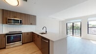 This single-floor unit showcases a contemporary design with a clean, open layout that connects the kitchen and living areas seamlessly. The kitchen is well-appointed with warm wood-tone cabinetry, white subway tile backsplash, a white quartz peninsula with an undermount sink and matte black faucet, and a full suite of stainless steel appliances including a range, microwave, and dishwasher. Light hardwood flooring extends throughout the main living areas, contributing to a bright and cohesive aesthetic.

The living area is spacious and sun-filled, anchored by expansive floor-to-ceiling sliding glass doors that open onto a private balcony with views of surrounding greenery, allowing abundant natural light to flood the interior. The neutral white walls and clean architectural lines enhance the airy atmosphere.

The bedroom is a comfortable, well-proportioned room featuring a large black-framed window that provides natural light and a closet for storage. The bathroom is finished with beige tile flooring, a tiled tub-shower surround, a wood-tone vanity, and a clean, functional layout. Throughout the unit, consistent finishes and a modern palette create a cohesive and polished living environment.